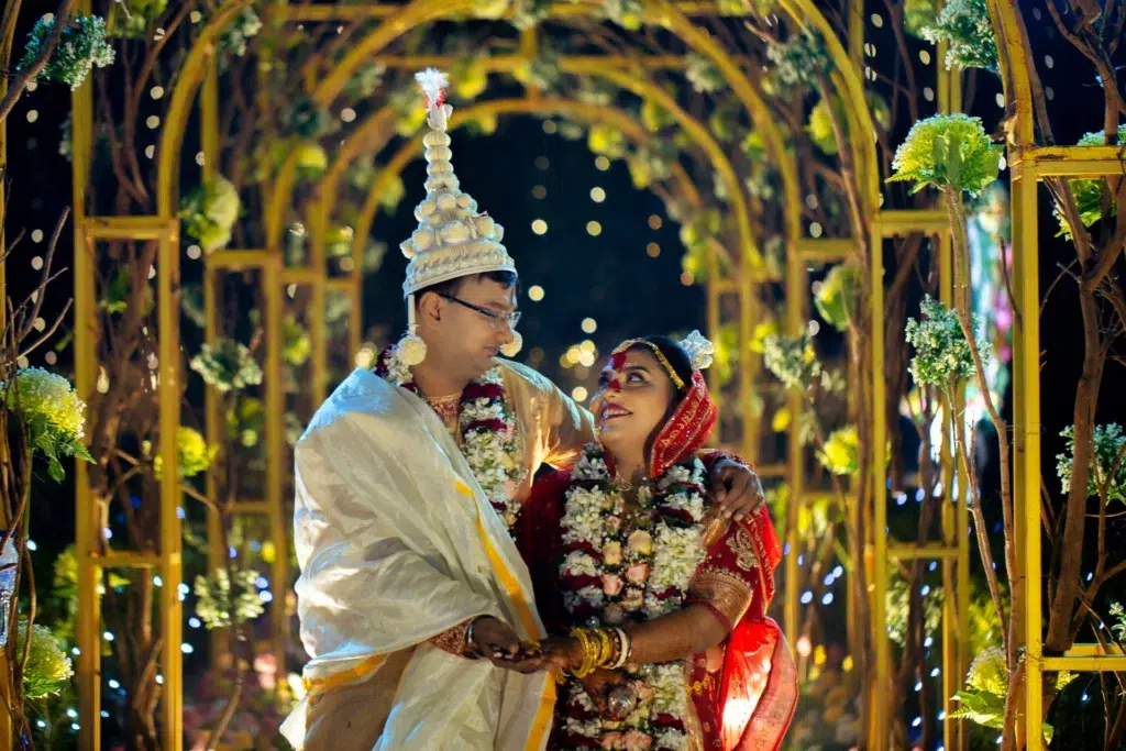 Bengali Bride And Groom Celebrating Under Golden Floral Trellis At Night — Groom In Dhoti, Bride In Saree With Traditional Headdress And Garlands At Wedding Ceremony In Kolkata By Sourav Deb